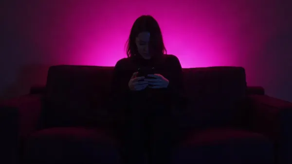 Silhouette seen from behind on a couch holding a phone, in a dark room with a magenta glow (article illustration)